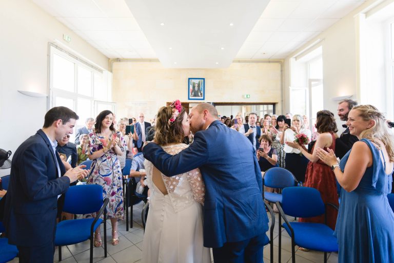 Aline et Yohan s’embrassent au centre de la salle des mariages de la mairie de Montussan, entourés d’invités assis aux tables, sous des guirlandes lumineuses chaleureuses et un grand cadre photo accroché au mur. ​