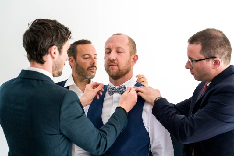 Quatre hommes en costume se tiennent serrés les uns contre les autres devant un fond blanc lumineux : au centre, le marié Yohan, les yeux fermés, porte un gilet et un nœud papillon bleus pendant que ses proches, souriants et concentrés, ajustent son nœud papillon et sa tenue, illustrant un moment complice de préparation juste avant la cérémonie.