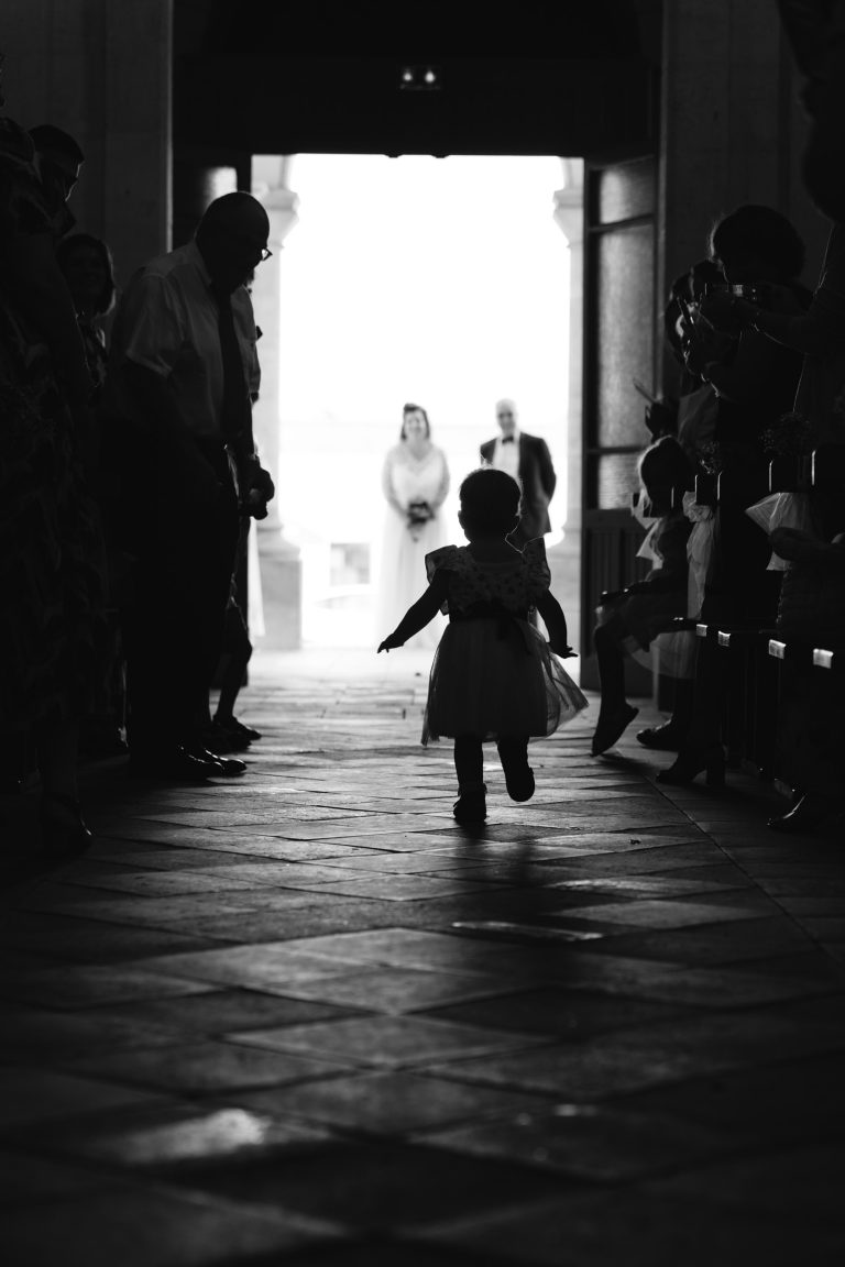 Un jeune enfant en robe blanche marche seul au centre de l’allée de l’église Saint‑Martin de Montussan, baignée de lumière par la grande porte ouverte au fond, tandis qu’Aline et Yohan se tiennent flous près de l’entrée et que les invités assis de part et d’autre observent la scène.