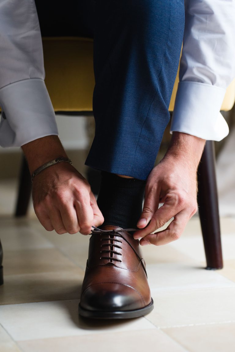 Le marié, Yohan, vêtu d’un pantalon bleu et d’une chemise blanche, est assis sur une chaise et se penche en avant pour nouer les lacets de ses chaussures de ville en cuir marron, la lumière naturelle mettant en valeur ses mains, la chaussure et le carrelage au sol juste avant la cérémonie. ​
