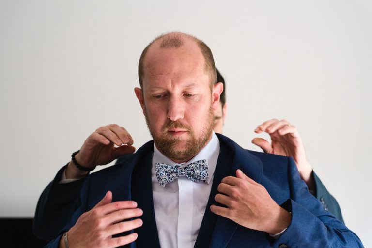 Le marié, Yohan, porte un costume bleu et un nœud papillon fleuri et se tient debout avec une expression concentrée tandis qu’une autre personne, partiellement cachée derrière lui, ajuste les épaules de sa veste sur un fond intérieur clair, juste avant la cérémonie de son mariage avec Aline.