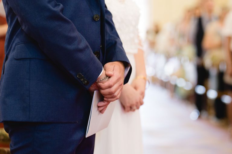 Yohan en costume bleu marine tient une enveloppe blanche entre ses mains jointes, debout à côté d’Aline en robe de mariée blanche, tous deux tournés vers des invités assis et flous au fond de l’église Saint‑Martin de Montussan pendant leur cérémonie. ​