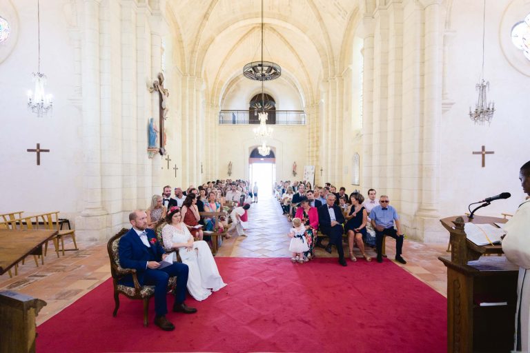 Vue depuis le fond de l’église Saint‑Martin de Montussan pendant la cérémonie de mariage d’Aline et Yohan, les mariés assis côte à côte devant l’autel éclairé par de grandes fenêtres, entourés de rangées d’invités et de lustres suspendus sous les hautes voûtes en pierre.