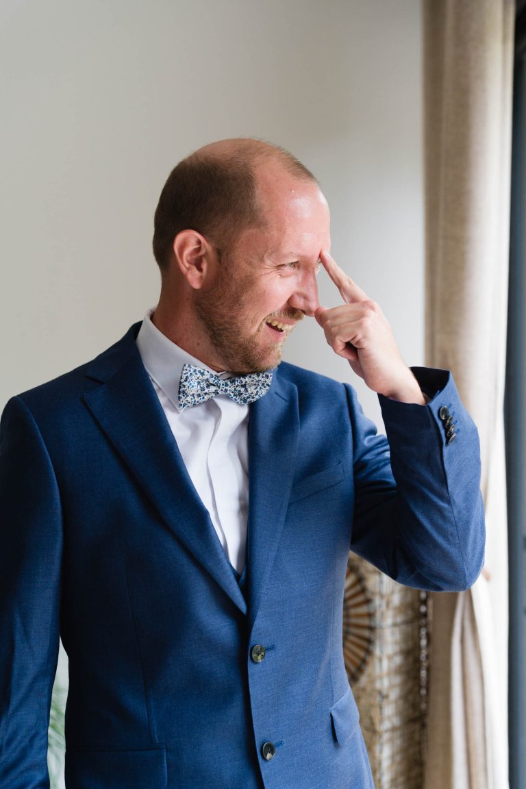 Le marié, Yohan, se tient à l’intérieur près d’une fenêtre aux rideaux beiges, vêtu d’un costume bleu, d’une chemise blanche et d’un nœud papillon à motifs, souriant en portant sa main droite à son front dans un moment de détente avant la cérémonie de son mariage avec Aline.