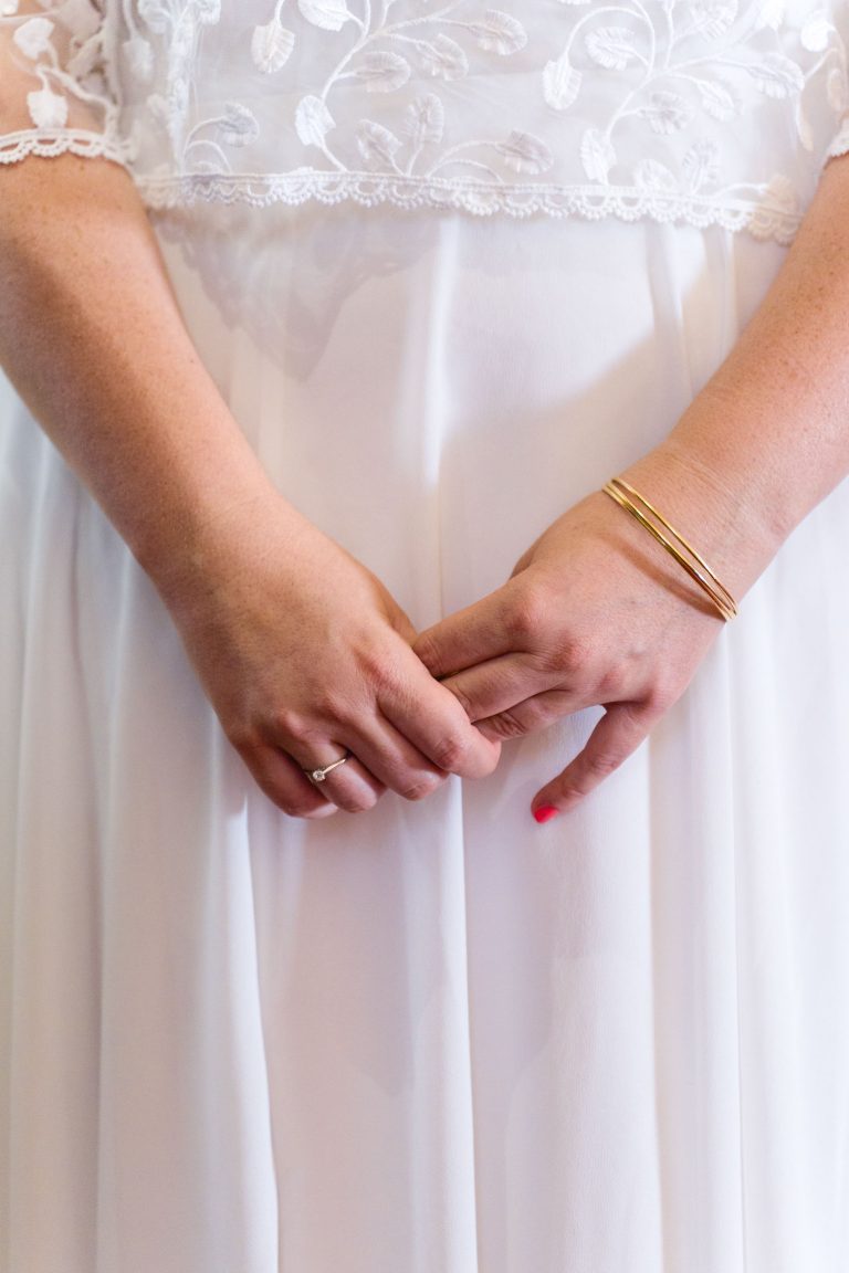 Gros plan sur la robe en dentelle blanche d’Aline, aux motifs floraux délicats, ses mains jointes à la taille portant un bracelet en or et une fine bague, avec des ongles courts dont l’un est verni en rose pastel.