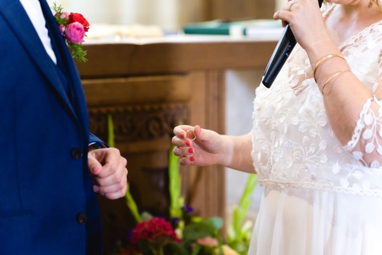 Aline en robe de mariée en dentelle tient un micro et prend la parole à l’église Saint‑Martin de Montussan, debout aux côtés de Yohan en costume bleu, devant un autel en bois décoré de bougies et de compositions florales colorées.