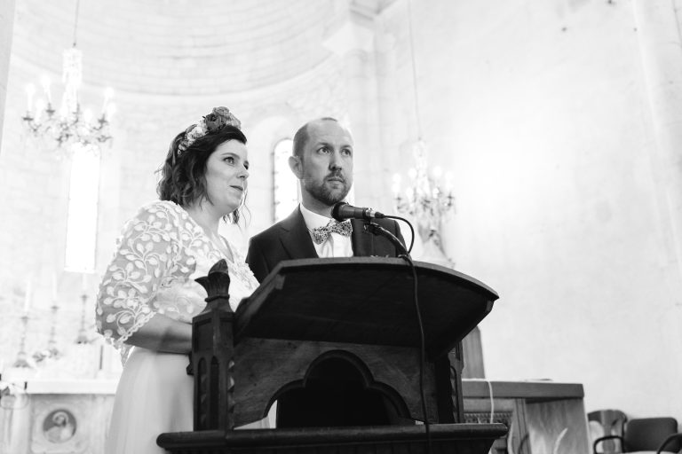 Aline en robe de mariée en dentelle et coiffe florale se tient aux côtés de Yohan en costume bleu sur l’estrade de l’église Saint-Martin de Montussan, tous deux souriants et debout derrière un pupitre avec micro, entourés de chandeliers dorés et de voûtes en pierre doucement éclairées.