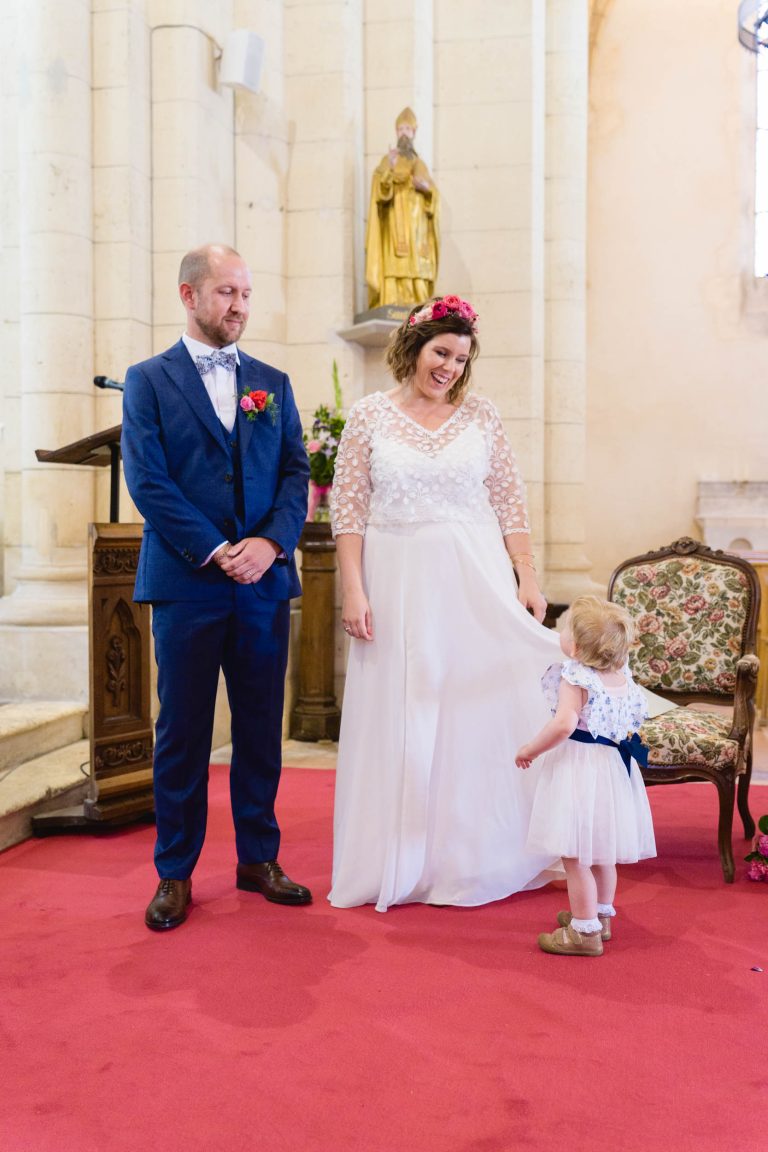 Aline en robe de mariée blanche et bouquet coloré se tient avec Yohan en costume bleu sur un tapis rouge à l’intérieur d’une église, souriant à une petite fille en robe blanche et sandales, entourés de chaises décorées de fleurs et d’une statue en arrière-plan.