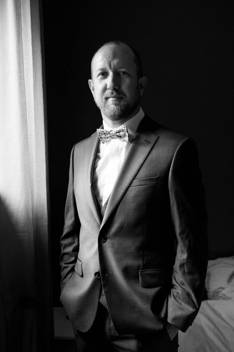 Texte alternatif optimisé : Yohan, le marié se tient à l’intérieur devant un fond sombre, vêtu d’un costume sombre, d’une chemise blanche et d’un nœud papillon à motifs, les mains dans les poches, tandis qu’une lumière douce venant d’une fenêtre à gauche éclaire son visage et crée une atmosphère élégante et intime
