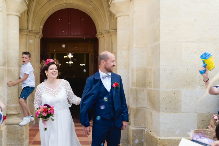 Aline en robe de mariée blanche et bouquet coloré au bras descend les marches d’un bâtiment en pierre aux côtés de Yohan en costume bleu, tous deux souriants, tandis que des invités soufflent des bulles de savon autour d’eux à la sortie de la cérémonie.