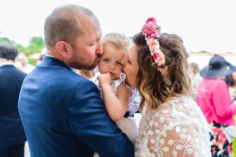 Aline en robe de mariée en dentelle et bandeau fleuri, accompagnée de Yohan en tenue de soirée, embrassent chacun une joue de leur fille habillé de blanc qui regarde l’objectif, tandis que l’arrière-plan extérieur lumineux reste flou.