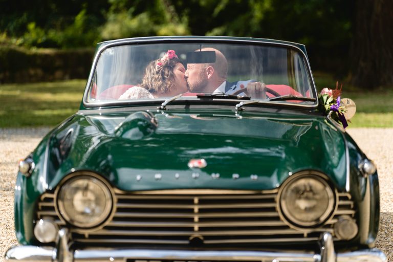 Aline et Yohan s’embrassent sur le siège avant d’une décapotable vintage verte garée sur un chemin de gravier, leurs silhouettes légèrement floues derrière le pare-brise sous une lumière de fin d’après-midi, entourés de verdure