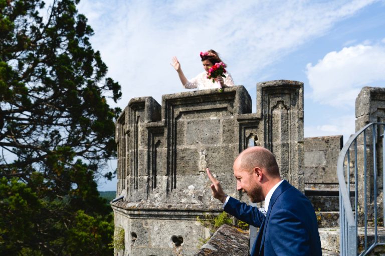 Aline en robe de mariée blanche et couronne de fleurs se tient sur la tourelle en pierre du Château de Camarsac et fait signe de la main à Yohan, en costume bleu, resté dans la cour au pied du château, sous un ciel bleu lumineux bordé d’un grand arbre vert.