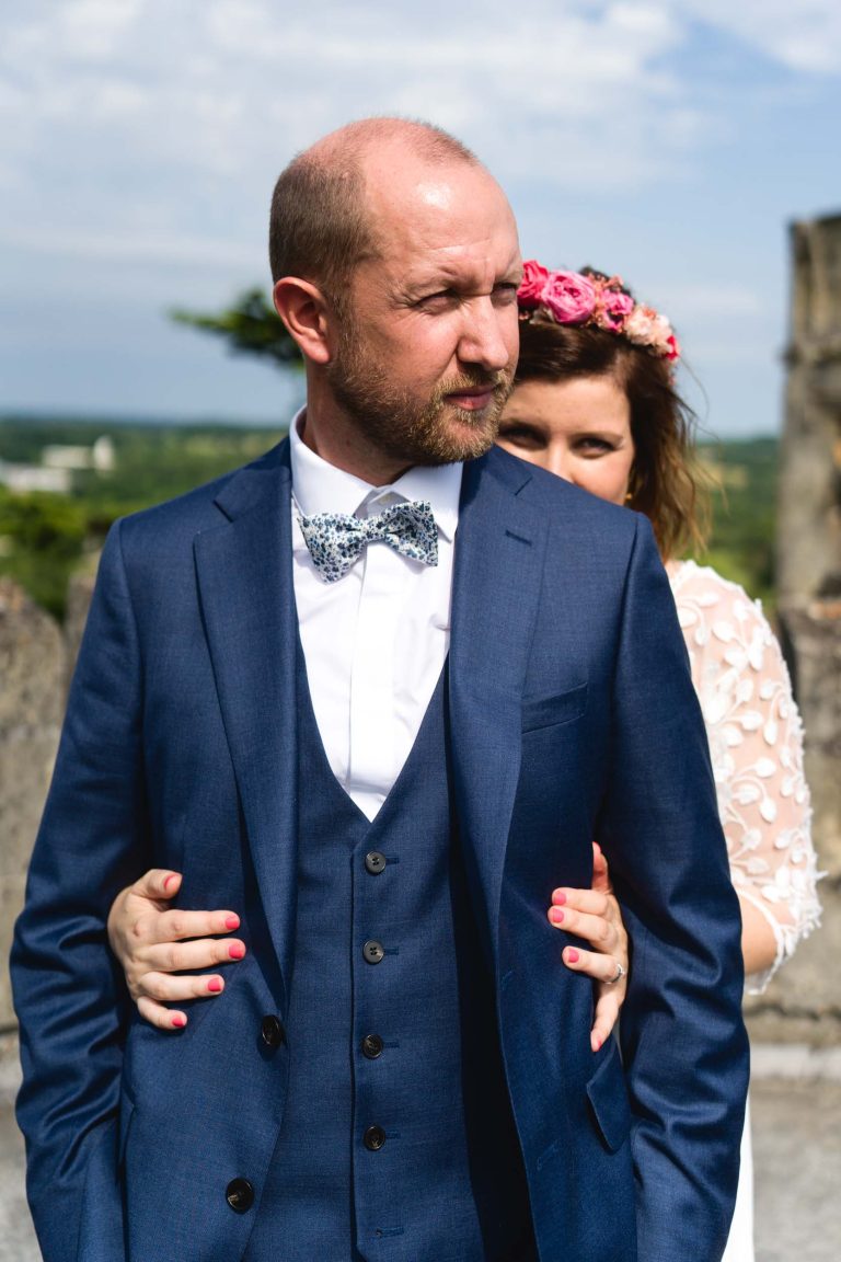 Yohan en costume bleu marine et nœud papillon à motifs regarde vers la droite, tandis qu’Aline en robe blanche et couronne de fleurs roses l’enlace par derrière, ses mains aux ongles vernis rose posées sur sa poitrine, sous une lumière extérieure douce et ensoleillée.