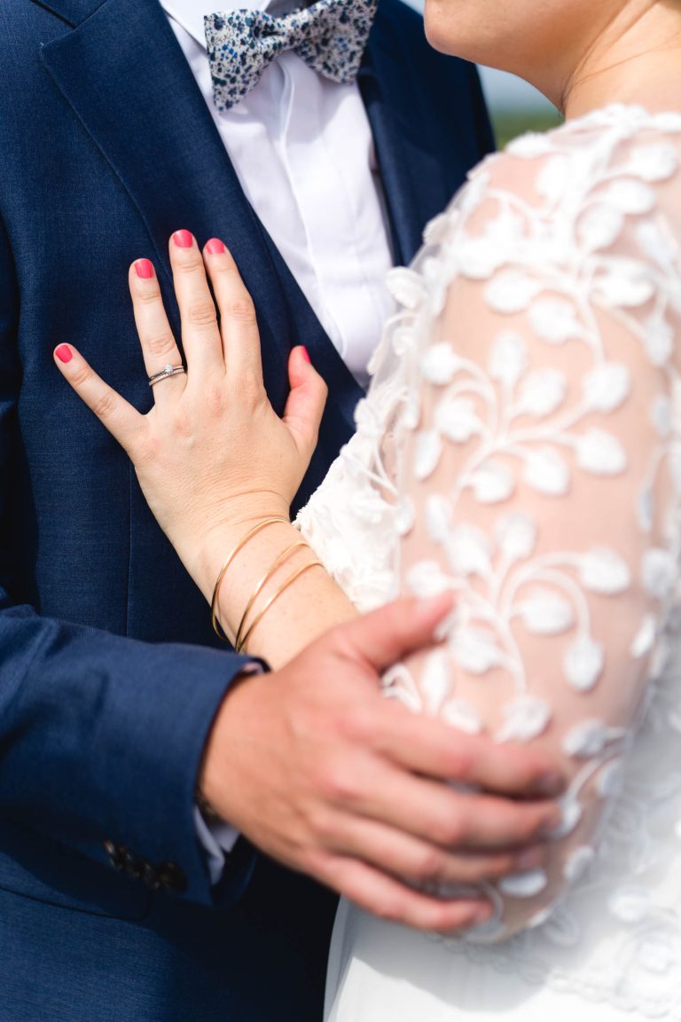 Gros plan sur Aline et Yohan enlacés : Aline en robe blanche aux manches en dentelle florale et aux ongles vernis rose pose sa main sur la poitrine de Yohan, vêtu d’un costume bleu marine avec nœud papillon à motifs, tandis qu’il tient délicatement son bras.