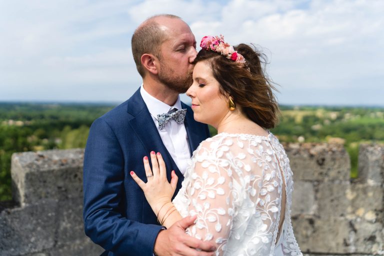 Yohan en costume bleu embrasse tendrement le front d’Aline en robe de mariée en dentelle blanche et coiffe florale, tous deux enlacés sur la terrasse en pierre du Château de Camarsac, avec en arrière-plan la campagne verdoyante et un ciel bleu légèrement nuageux.