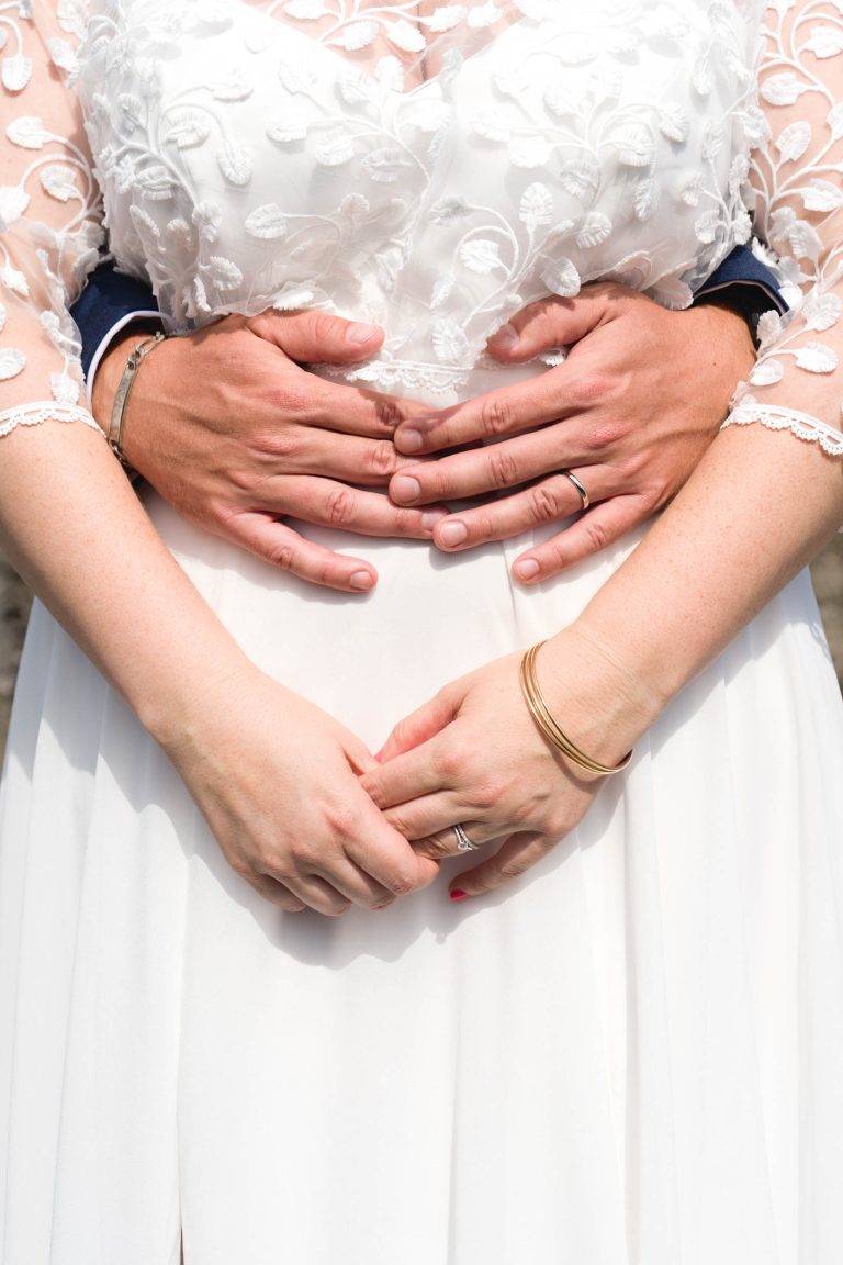 Gros plan sur les mains d’Aline et de Yohan en tenue de mariage qui s’enlacent pendant un baiser, Aline en robe blanche brodée au premier plan, Yohan en costume sombre à l’arrière-plan, tous deux portant leurs alliances et des bracelets en or, leurs visages restant hors champ pour concentrer l’attention sur le geste tendre