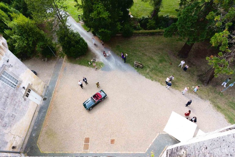 Vue aérienne de la cour intérieure du Château de Camarsac, avec une voiture décapotable verte garée sur le gravier au centre, entourée d’invités en tenues de mariage colorées qui circulent et discutent près des arbres, des massifs de verdure et des façades en pierre claire du domaine