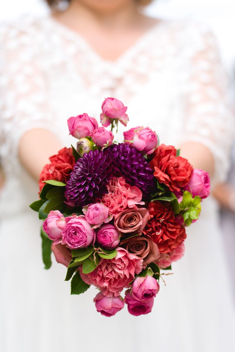 Une mariée en robe en dentelle blanche tient son bouquet coloré de roses roses, d’œillets rouges, de dahlias violets et de verdure, présenté en gros plan devant l’objectif lors du mariage d’Aline et Yohan