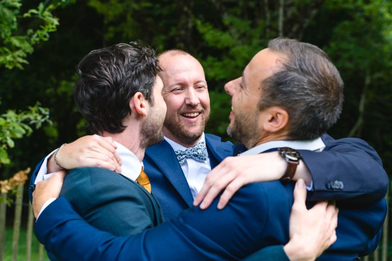 Trois hommes en costume se tiennent enlacés en extérieur, se serrant l’un contre l’autre dans une étreinte chaleureuse et joyeuse, tous souriant et riant au milieu d’un décor de verdure légèrement flou, illustrant un moment complice du mariage d’Aline et Yohan.
