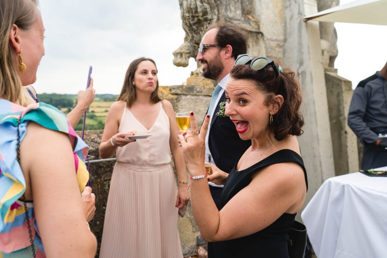 Une femme souriante, vêtue d'une robe noire et portant des lunettes de soleil, fait des gestes ludiques vers la caméra tandis que d'autres personnes discutent derrière elle lors d'un événement en plein air. Avec un style vibrant, des collines verdoyantes s'étendent à l'arrière-plan.