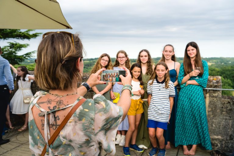 Une femme aux cheveux courts et à la robe fleurie prend une photo avec son téléphone de huit filles souriantes en tenue d'été colorée, debout en plein air sur une terrasse en pierre sous un ciel nuageux. Le paysage luxuriant constitue une toile de fond parfaite.