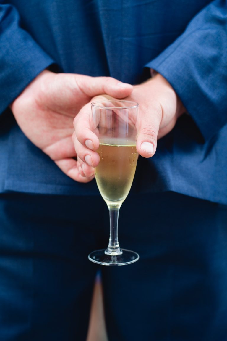 Une personne en costume bleu tient à deux mains une coupe de champagne derrière son dos, la boisson légère et pétillante attirant le regard. Seuls les mains, le verre et le bas de la veste du costume sont visibles dans l'image, ce qui donne une touche d'élégance.