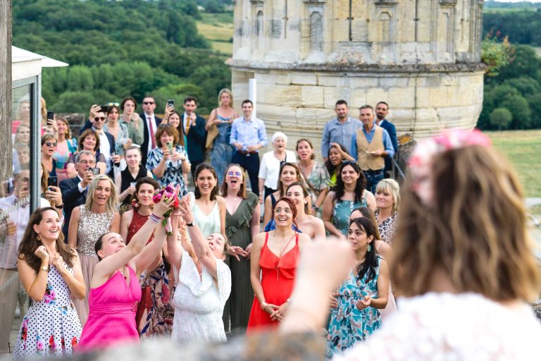 Lors d'un mariage, un groupe de femmes, vêtues de robes d'été colorées, tendent la main pour attraper un bouquet. Les invités derrière elles sourient et regardent. La scène se déroule à l'extérieur, avec de la verdure, un vieux bâtiment en pierre et une mariée floue au premier plan.