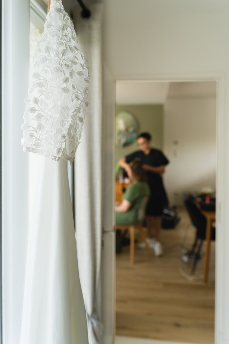 Une robe de mariée blanche en dentelle florale est suspendue près d’une fenêtre dans une chambre lumineuse à Montussan, tandis qu’en arrière‑plan flou une coiffeuse prépare une invitée, avec parquet en bois, miroir rond et décoration douce créant une atmosphère élégante de préparation de mariage.