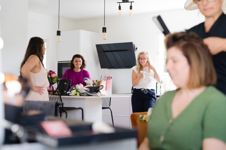 Quatre femmes se trouvent dans une cuisine moderne et lumineuse à Montussan : au premier plan, une invitée en robe verte se fait coiffer, légèrement floue, tandis qu’à l’arrière‑plan trois femmes debout discutent et sourient près de l’îlot de cuisine, entourées de sacs et de produits de maquillage, dans une ambiance conviviale de préparation de mariage. ​