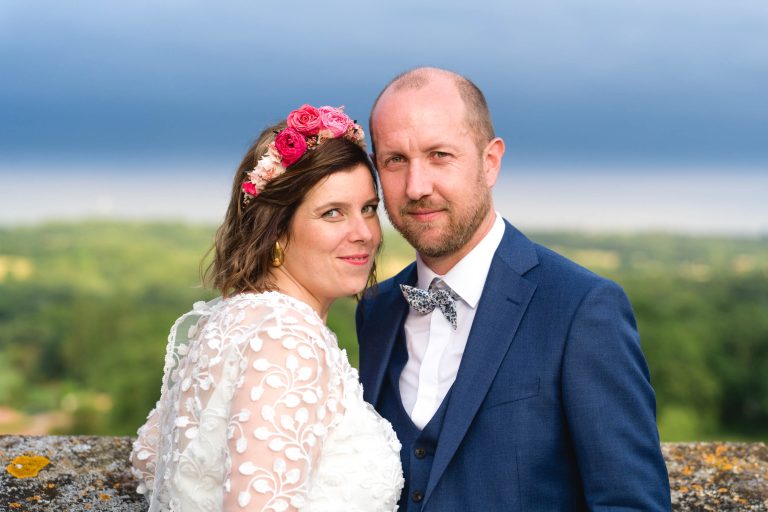 Une femme vêtue d'une robe en dentelle blanche et d'une couronne de fleurs roses se tient près d'un homme en costume bleu et nœud papillon fleuri. Ils regardent l'appareil photo avec un doux sourire, dans un paysage pittoresque et verdoyant sous un ciel bleu foncé.