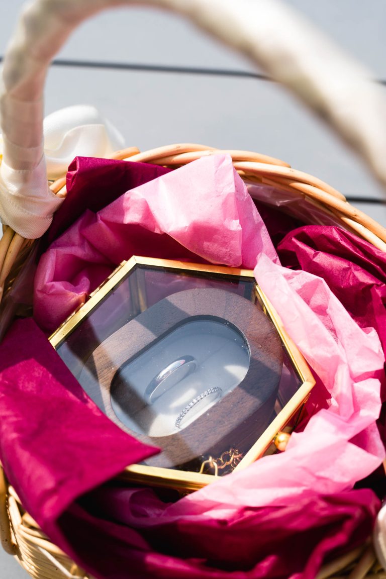 Deux alliances en argent reposent dans une petite boîte en verre posée dans un panier en osier tapissé de papier de soie rose et bordeaux, éclairées par une lumière naturelle douce qui met en valeur les textures et crée une ambiance élégante et festive pour le mariage d’Aline et Yohan. ​