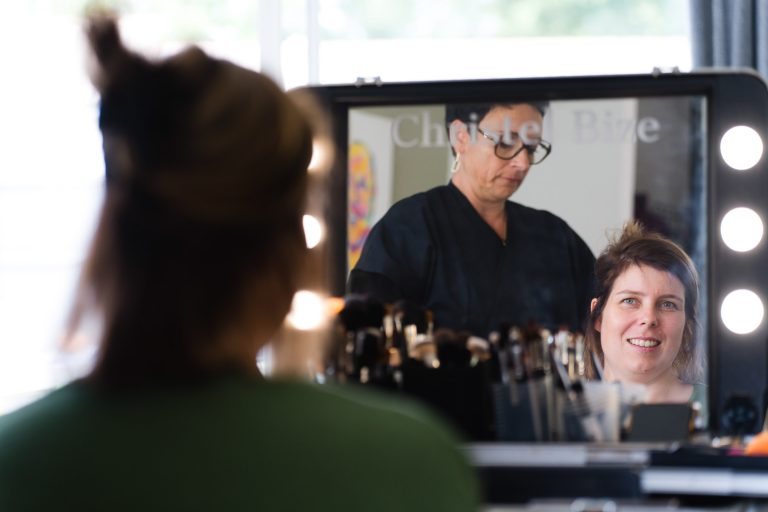 Aline assise devant une coiffeuse lumineuse à Montussan sourit à son reflet pendant que Chrystelle bize derrière elle ajuste délicatement ses cheveux, des pinceaux et produits de maquillage étant disposés sur la table dans une atmosphère chaleureuse de préparation de mariage.