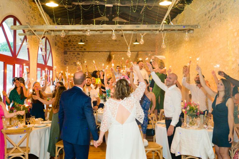 Les mariés, vus de dos, entrent main dans la main dans une salle rustique et chaleureusement éclairée, magnifiquement stylisée, et remplie d'invités qui les acclament. Les murs en pierre, les poutres en bois et les lumières suspendues créent une atmosphère de fête étonnante.