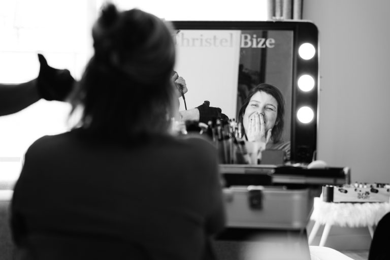 Aline assise de dos devant une coiffeuse lumineuse à Montussan sourit en se couvrant la bouche de surprise ou de joie en regardant son reflet dans un miroir entouré d’ampoules, tandis que des pinceaux et palettes de maquillage sont disposés sur la table et qu’une main gantée apparaît à côté d’elle, illustrant un moment intime de préparation de mariage.