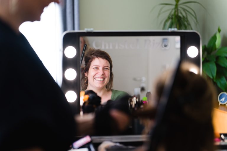 Une femme aux cheveux bruns courts, assise devant une coiffeuse dans un salon lumineux à Montussan, sourit chaleureusement à son reflet dans un miroir entouré de lumières, tandis qu’une styliste floue s’affaire à l’arrière‑plan près d’outils de maquillage et d’une plante verte.