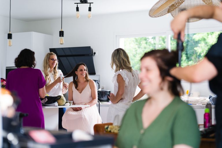 Cinq femmes sont réunies dans une cuisine moderne et lumineuse à Montussan : une femme en blanc, assise, sourit à une proche debout à ses côtés, tandis qu’au fond deux femmes échangent près du plan de travail et qu’une coiffeuse boucle les cheveux d’une invitée en vert, dans une ambiance animée et détendue de préparation de mariage.