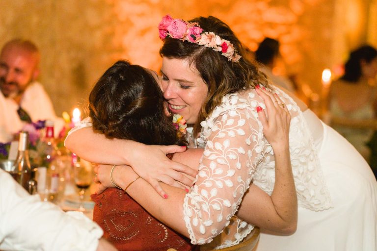 Une mariée souriante, vêtue d'une robe blanche avec des manches en dentelle florale et une couronne de fleurs roses, étreint chaleureusement un invité lors d'une réception aux chandelles. Cette scène joyeuse se caractérise par des invités légèrement flous et un éclairage chaleureux en arrière-plan.