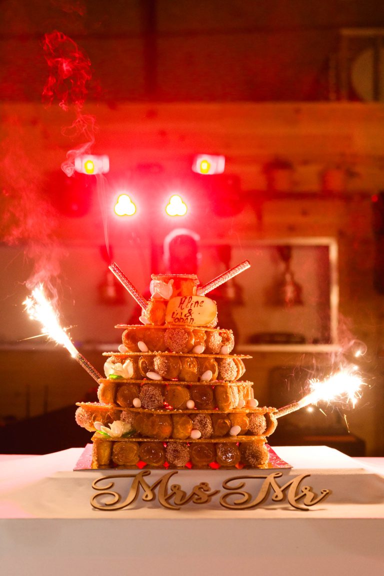 Un croquembouche à étages décoré de cierges magiques, de bâtonnets de gaufrettes et d'une plaque "Pline et Chloé" est posé sur une table avec un panneau "Mme et M." en or, tandis que des lumières rouges illuminent l'arrière-plan.