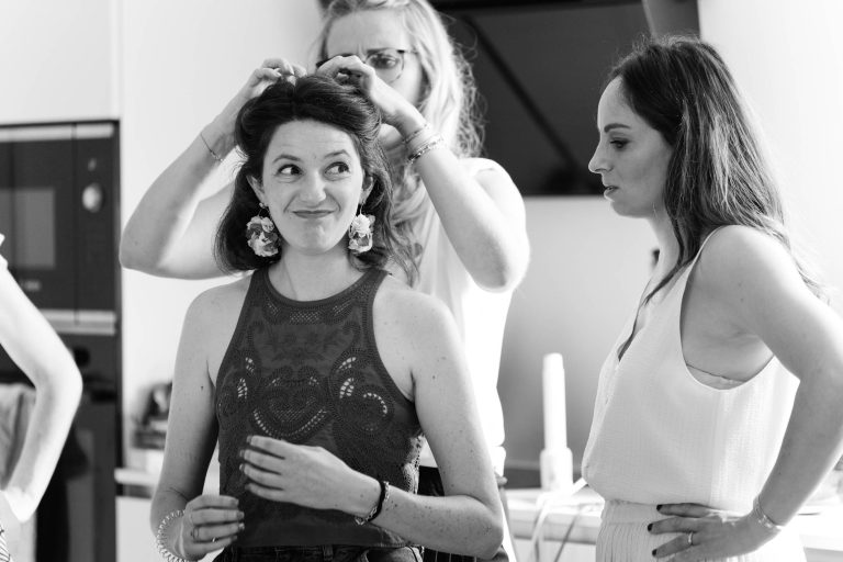 Trois femmes se tiennent dans une pièce moderne et lumineuse à Montussan : au centre, une jeune femme souriante en robe claire se fait coiffer par une professionnelle en noir qui lui boucle les cheveux, tandis qu’une troisième femme l’observe avec attention à l’arrière‑plan, dans une scène de préparation de mariage en noir et blanc.