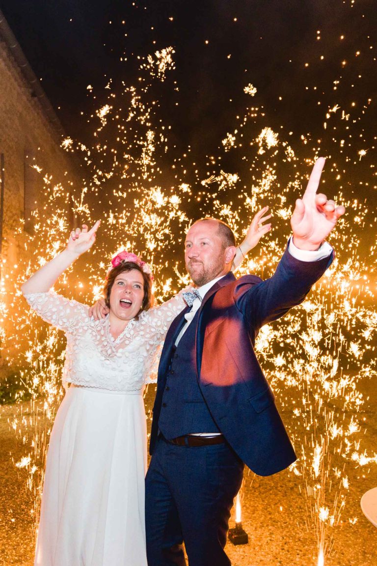 Un couple joyeux en tenue de mariage fait la fête en plein air la nuit, les bras levés et le sourire aux lèvres devant des cierges magiques, créant ainsi une atmosphère énergique et festive.