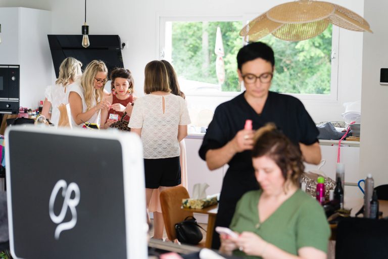 Dans une grande pièce moderne et lumineuse à Montussan, plusieurs femmes se préparent pour le mariage d’Aline : au premier plan une coiffeuse en noir boucle les cheveux d’une invitée assise, tandis qu’au fond d’autres proches discutent, se maquillent et grignotent près de la fenêtre, dans une ambiance conviviale et animée. ​