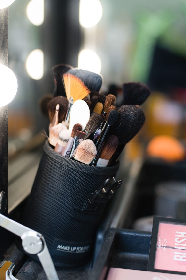 Un pot cylindrique noir rempli de pinceaux de maquillage, de peignes et d’outils de coiffure est posé sur une coiffeuse blanche bien éclairée, entouré de flacons et produits flous en arrière‑plan, créant une atmosphère professionnelle de préparation de mariée à Montussan. ​