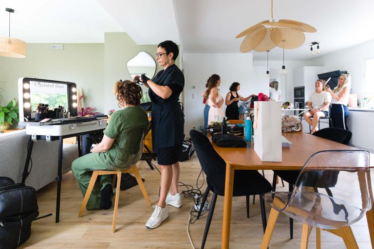 Chrystelle Bize en robe noire boucle les cheveux d’Aline assise dans une pièce moderne et lumineuse à Montussan, tandis qu’au second plan plusieurs proches discutent et se préparent autour de tables couvertes de matériels de coiffure et de maquillage, sur un parquet en bois clair éclairé par de grandes fenêtres et des lampes suspendues.