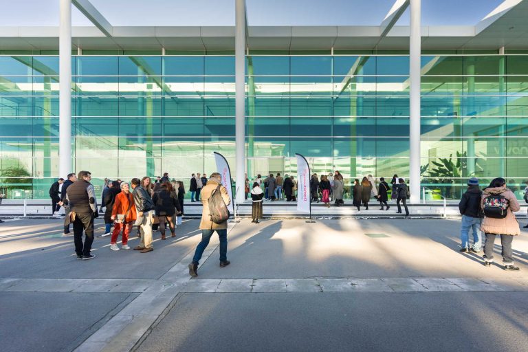 Un groupe de personnes se tient debout et marche à l'extérieur d'un bâtiment moderne DOMOFRANCE en verre avec des piliers blancs par une journée ensoleillée. Certains tiennent des sacs ou portent des manteaux. Deux drapeaux blancs sont installés près de l'entrée, se reflétant dans les grandes fenêtres en verre.