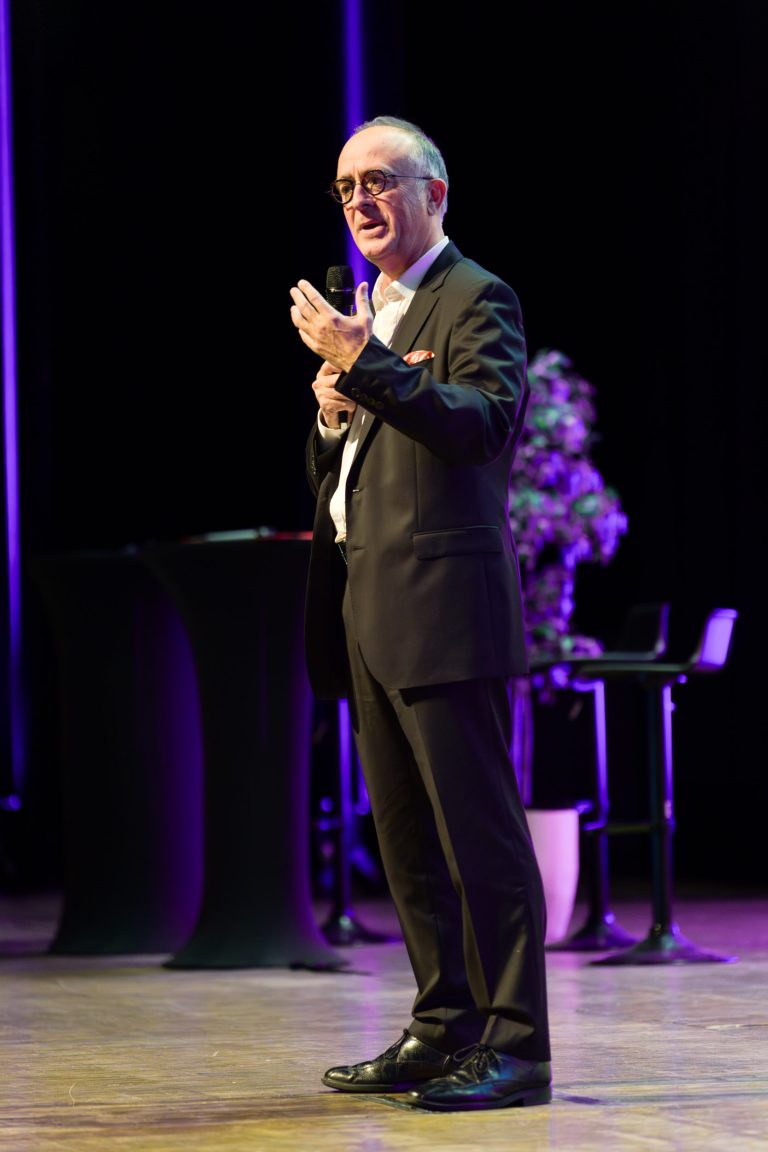 Un homme en costume sombre se tient sur une scène et parle dans un microphone lors de l'événement DOMOFRANCE. Il fait des gestes d'une main, semblant engagé et expressif sur un fond violet faiblement éclairé, avec une composition florale et des chaises visibles derrière lui.