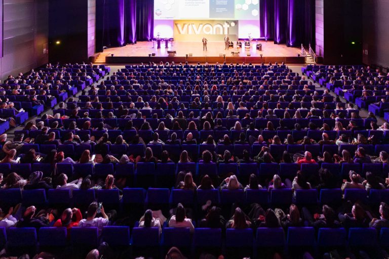 Un public nombreux est assis sur des rangées de sièges bleus, face à une scène très éclairée où se tiennent plusieurs orateurs. Derrière eux, un écran affiche des diapositives et le mot "Vivant", mettant en évidence DOMOFRANCE. L'auditorium spacieux est doté d'un éclairage violet et d'un décor moderne.