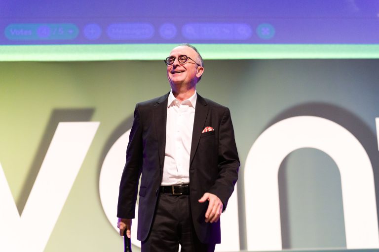 Un homme souriant, vêtu d'un costume noir, d'une chemise blanche et de lunettes, se tient avec assurance sur la scène devant de grandes lettres blanches DOMOFRANCE. L'arrière-plan est éclairé par des teintes vertes et violettes, et il tient une canne dans sa main droite.