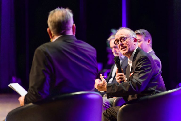 Un homme en costume tenant un micro parle avec animation sur scène lors d'une table ronde de DOMOFRANCE. Trois autres hommes en costume sont assis à ses côtés, l'un d'entre eux étant au premier plan et faisant face à la scène. La scène est éclairée par des lumières violettes vibrantes.