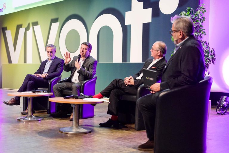 Quatre hommes en costume sont assis sur une scène, sur des chaises noires, et participent à une table ronde. L'un d'eux fait des gestes pendant qu'il parle et les autres écoutent attentivement. En arrière-plan, un grand panneau "DOMOFRANCE vivant" et une plante en pot près de la table ronde.
